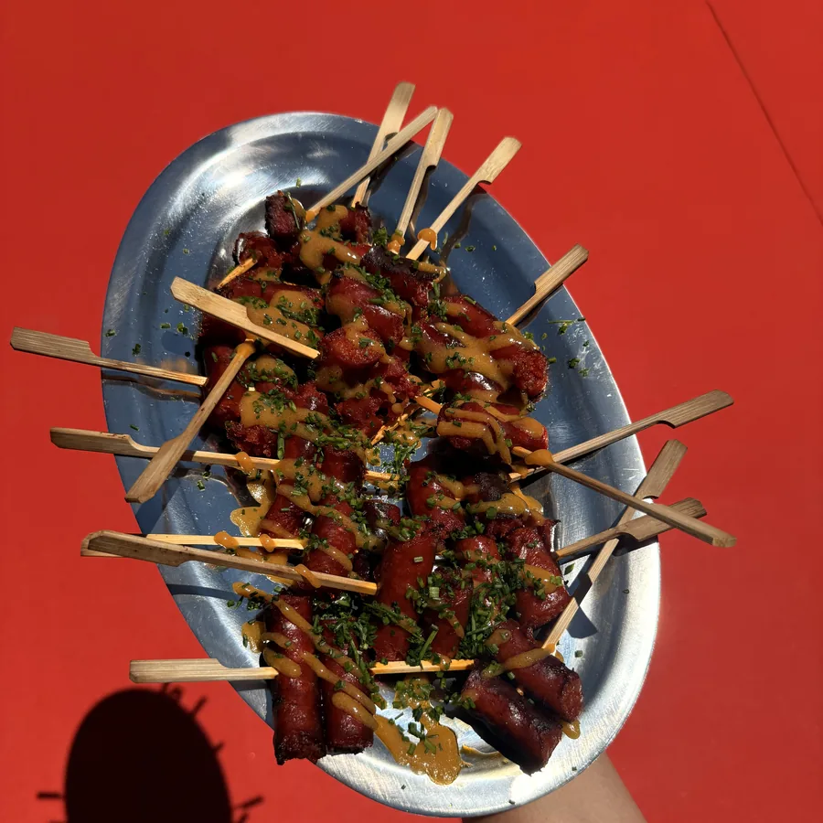 Bandeja metálica con pinchos de chistorra, salsa y cebollino picado, sobre un fondo rojo