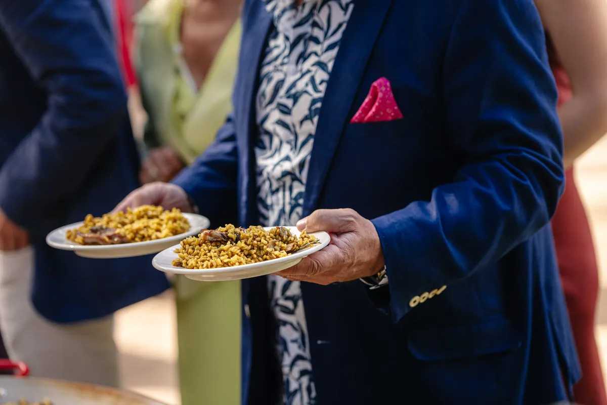 Un invitado con chaqueta azul y pañuelo rosa en el bolsillo lleva dos platos de arroz recién servido