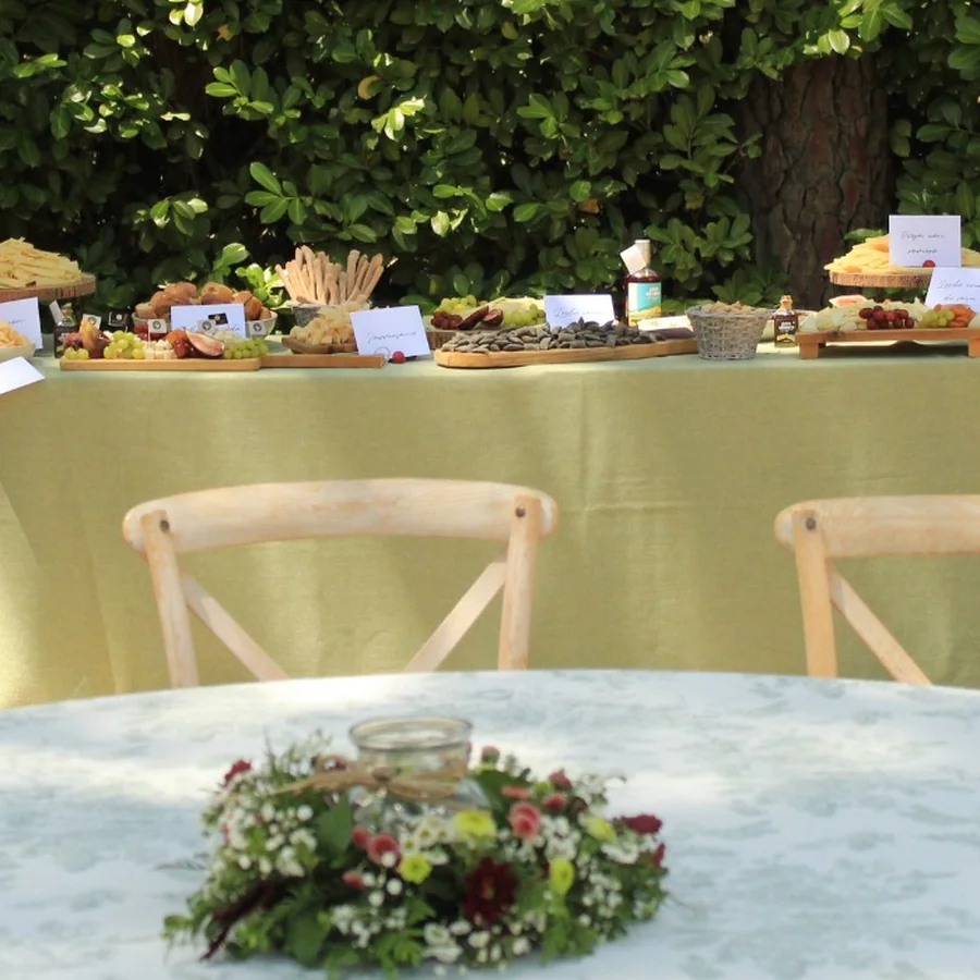 Mesa redonda con mantel claro y un centro de flores en un jardín; detrás, una mesa larga con tablas de queso, uvas y carteles