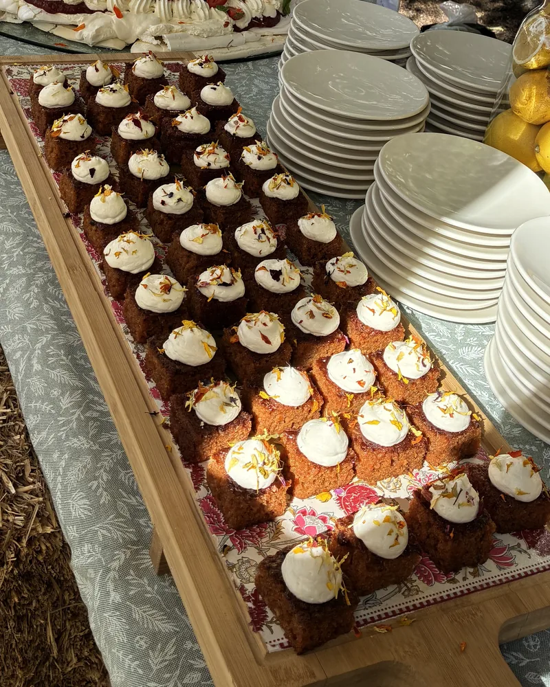 Tabla de madera con bizcochitos cuadrados coronados de crema y pétalos, junto a pilas de platos blancos