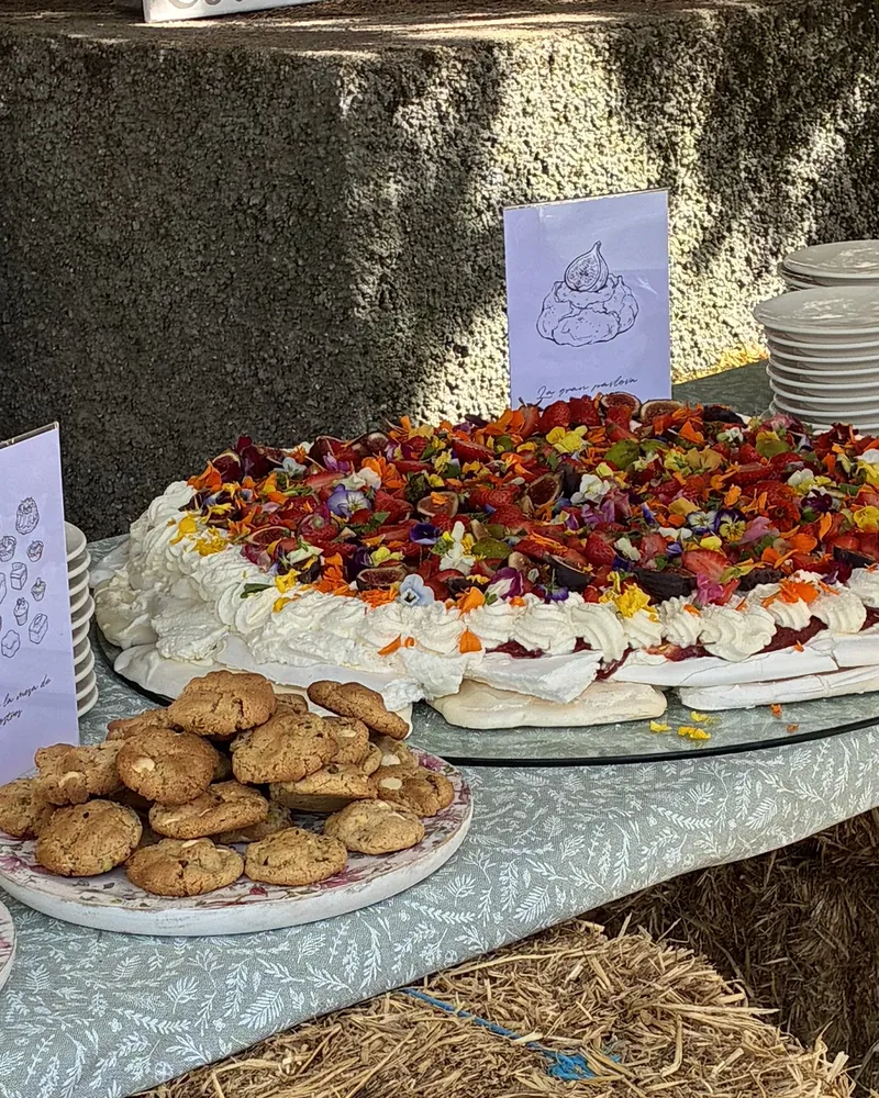 Tarta de merengue cubierta de fruta y flores, con un plato de galletas delante, en una Mesa de Postres al aire libre